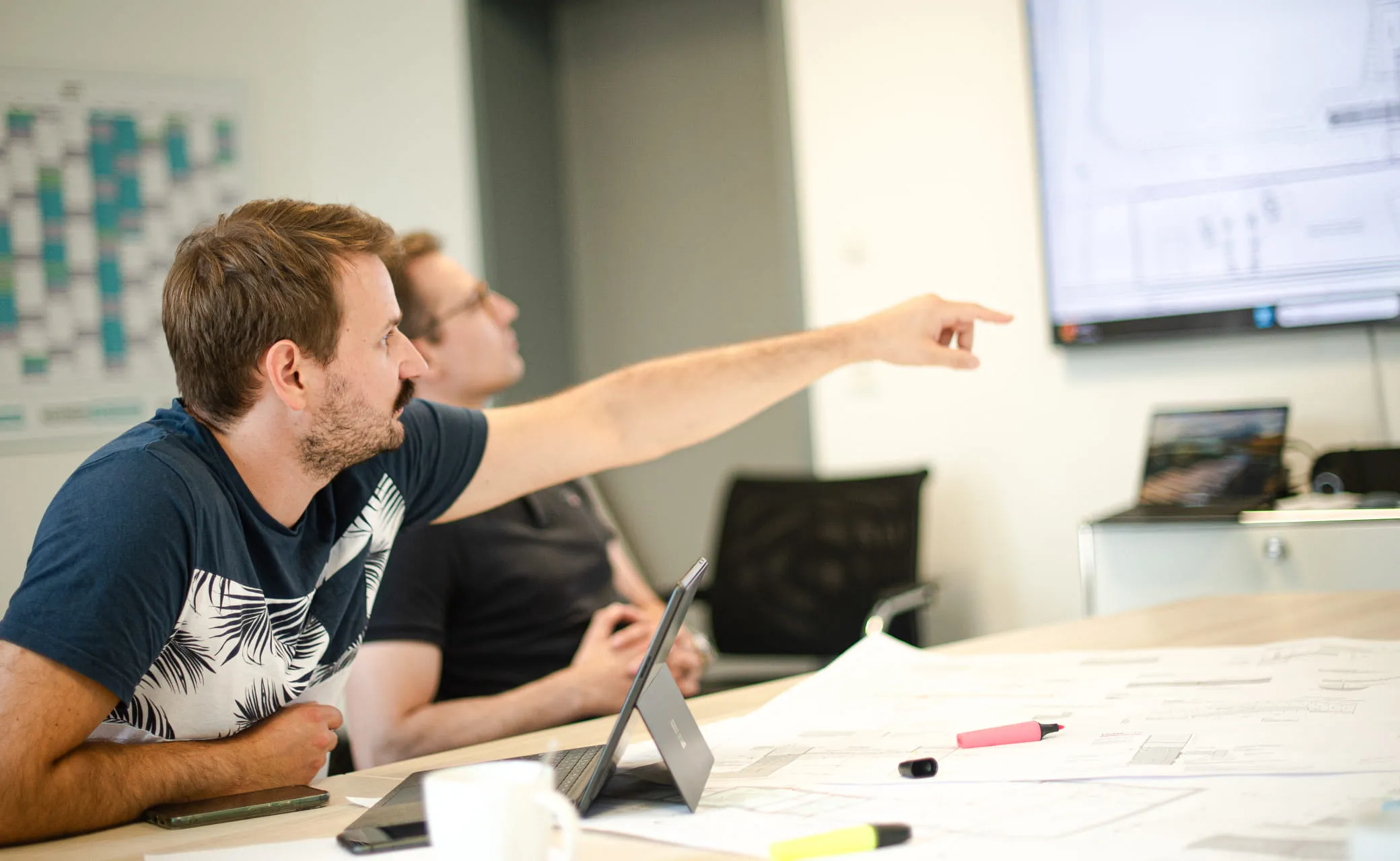 Zwei Mitarbeiter an Tisch sitzend, davon einer auf Bildschirm im Hintergrund deutend